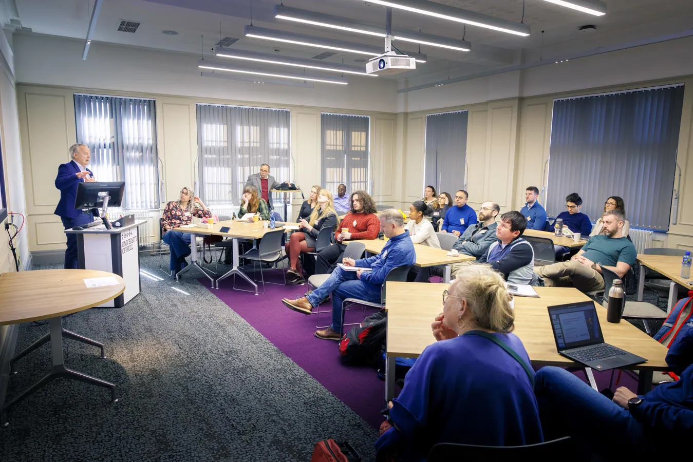 Groups of people sit around tables listening to a speaker delivering a presentation