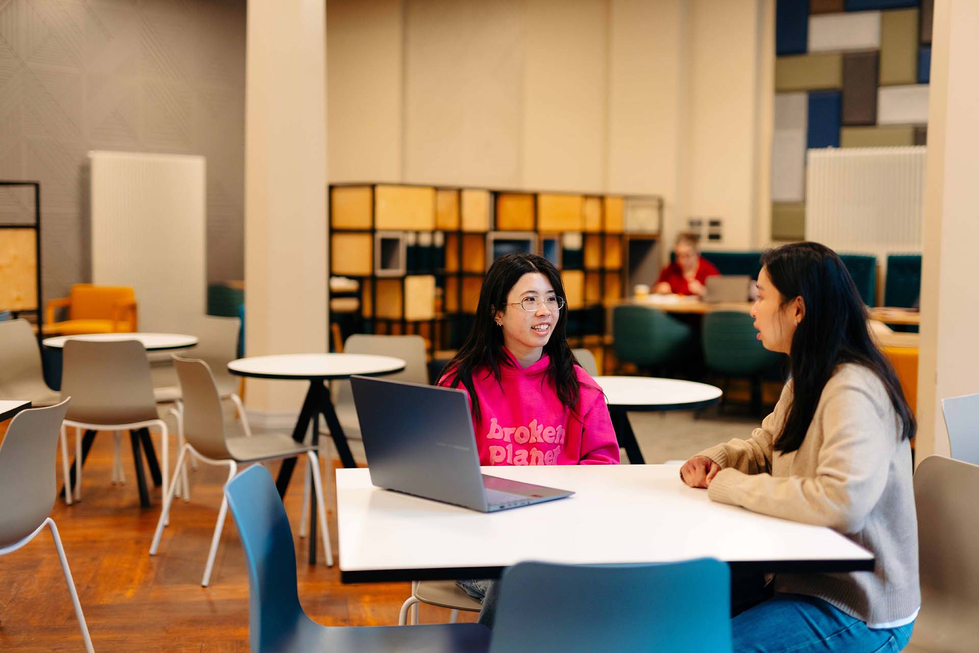 Two women sit chatting at a table over a laptop