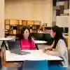 Two women sit chatting at a table over a laptop