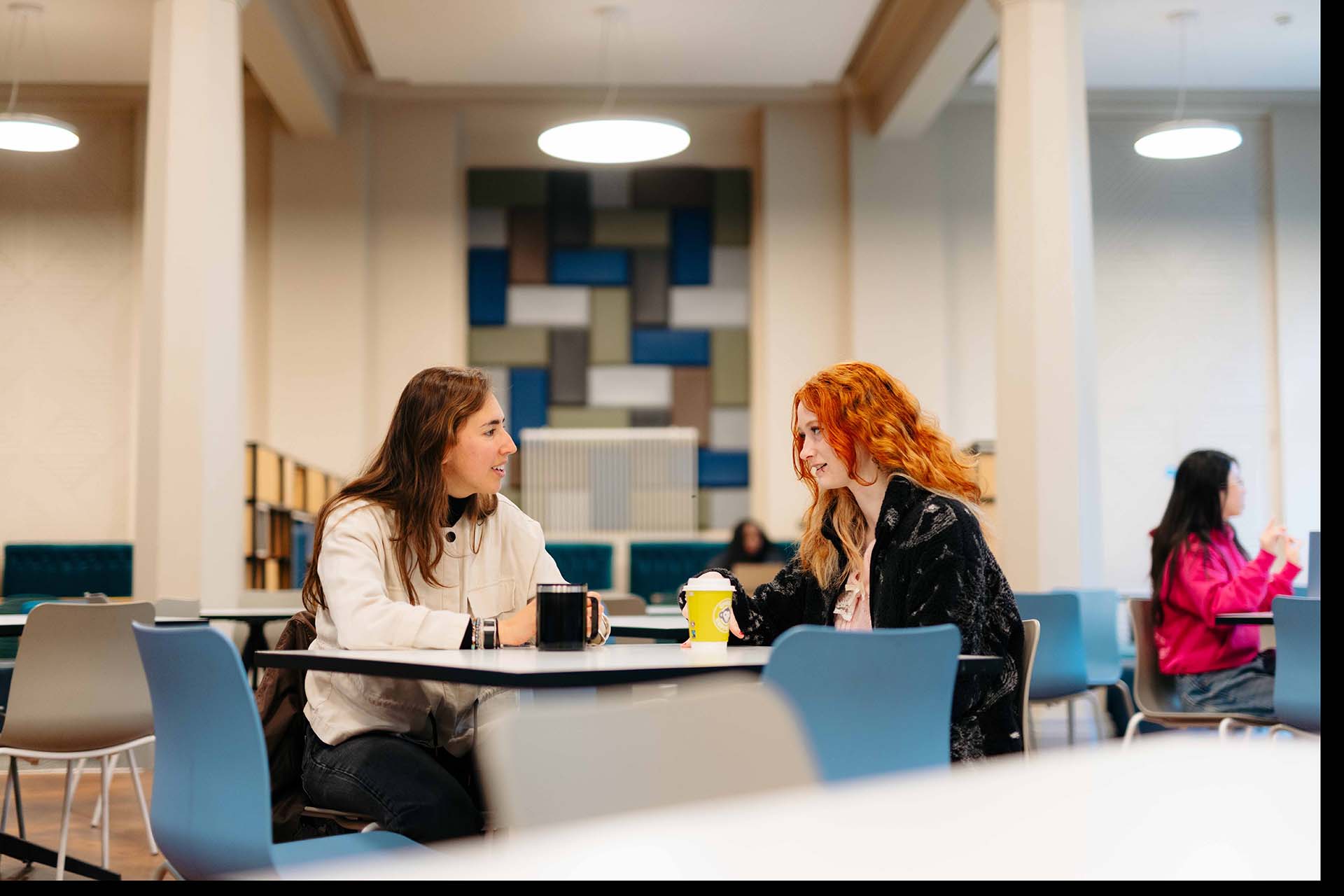 Two women sit chatting at a table over coffees