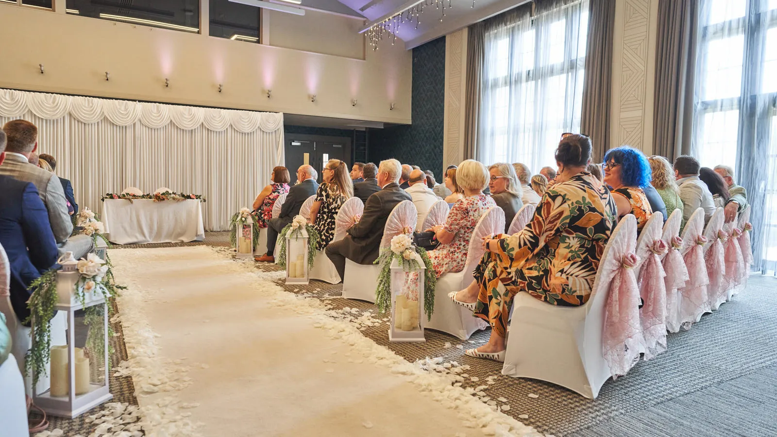 Wedding guests seated in rows next to a white aisle runner inside a large conference-style reception room.