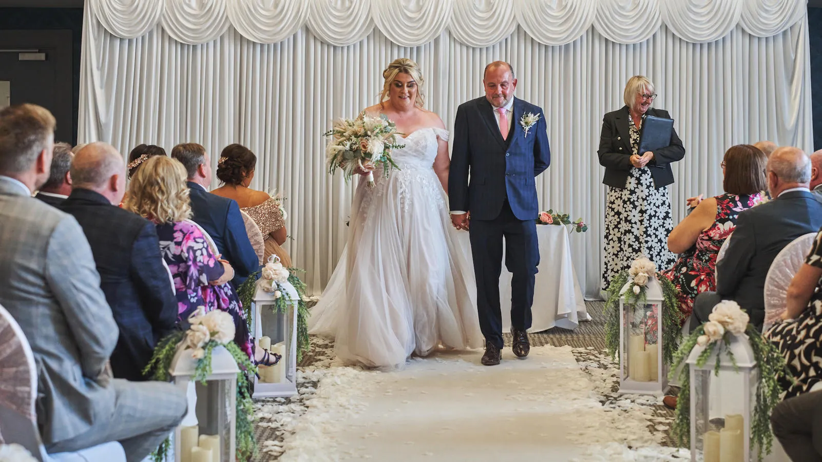 A bride and groom standing in front of a white backdrop at the end of a wedding aisle.
