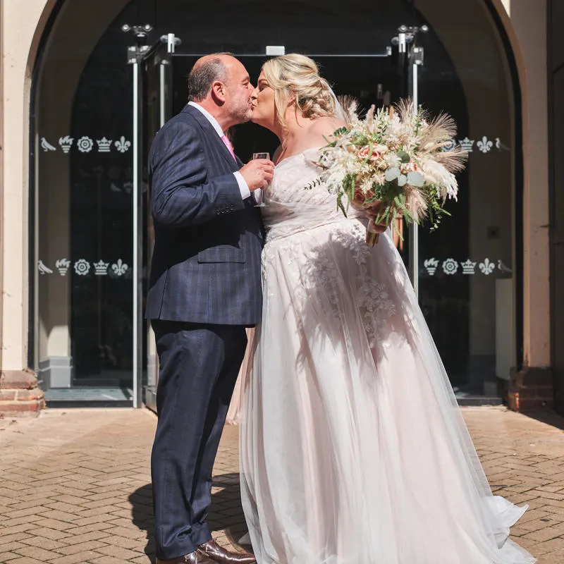 Bride and groom sharing a kiss outside their wedding venue, celebrating with champagne in the sunshine.
