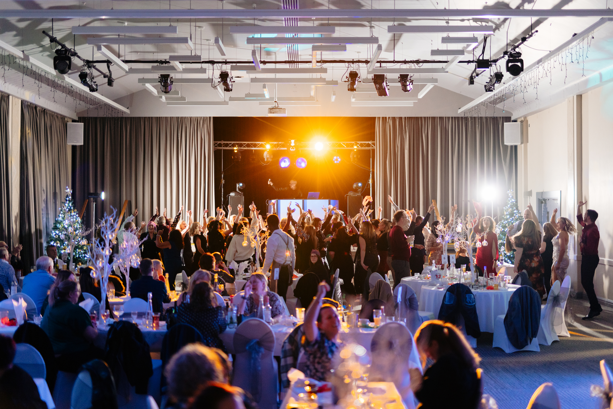 A large group of people dancing under coloured lights in front of a DJ booth in a large function room