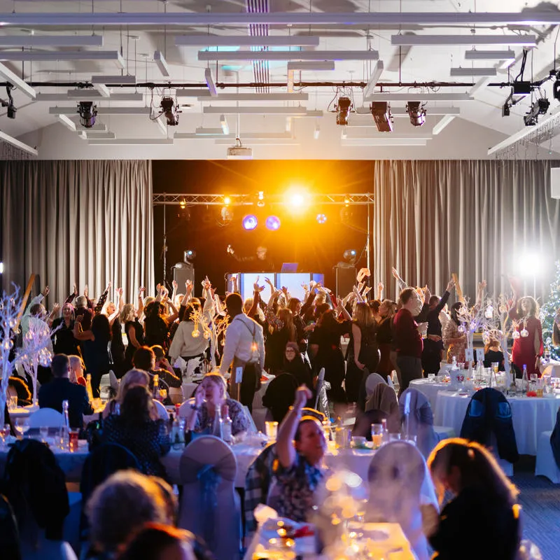A large group of people dancing under coloured lights in front of a DJ booth in a large function room
